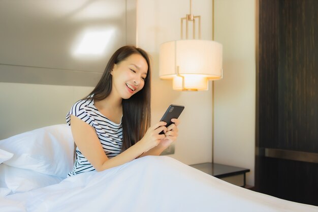 La bella giovane donna asiatica del ritratto utilizza il telefono cellulare astuto sul letto nell'interno della camera da letto