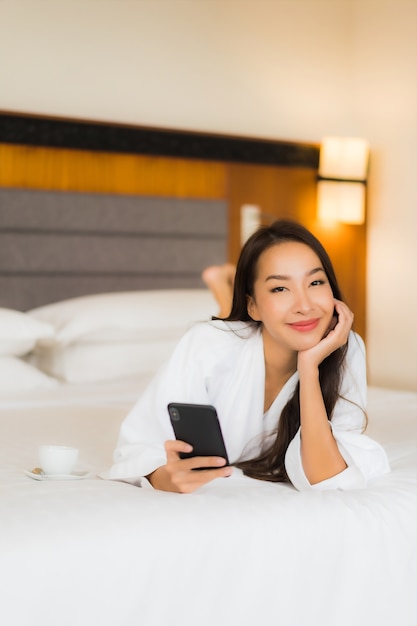 La bella giovane donna asiatica del ritratto utilizza il telefono cellulare astuto sul letto nell'interno della camera da letto