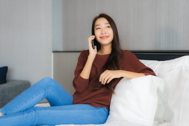 La bella giovane donna asiatica del ritratto utilizza il telefono cellulare astuto sul letto nell'interno della camera da letto