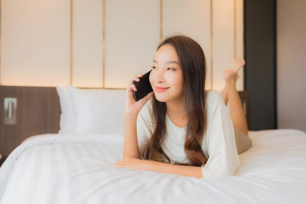 La bella giovane donna asiatica del ritratto utilizza il telefono cellulare astuto sul letto nell'interno della camera da letto