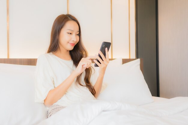 La bella giovane donna asiatica del ritratto utilizza il telefono cellulare astuto sul letto nell'interno della camera da letto