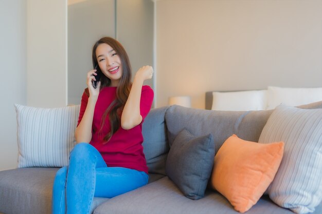 La bella giovane donna asiatica del ritratto utilizza il telefono cellulare astuto sul sofà