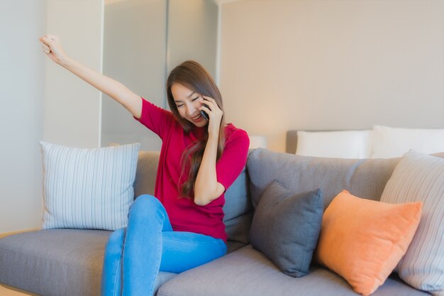 La bella giovane donna asiatica del ritratto utilizza il telefono cellulare astuto sul sofà