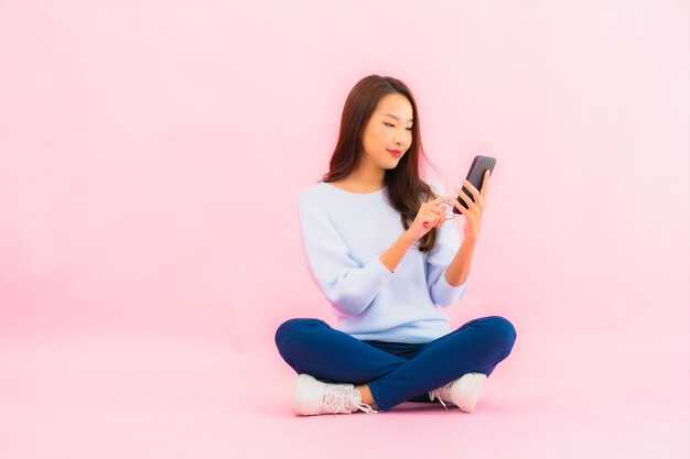 La bella giovane donna asiatica del ritratto utilizza il telefono cellulare astuto sulla parete isolata di colore rosa