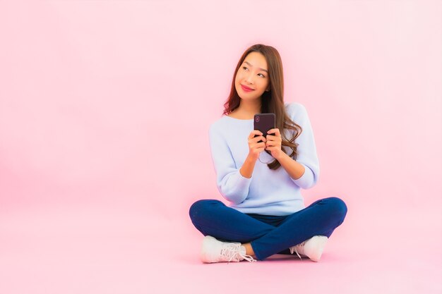 La bella giovane donna asiatica del ritratto utilizza il telefono cellulare astuto sulla parete isolata di colore rosa