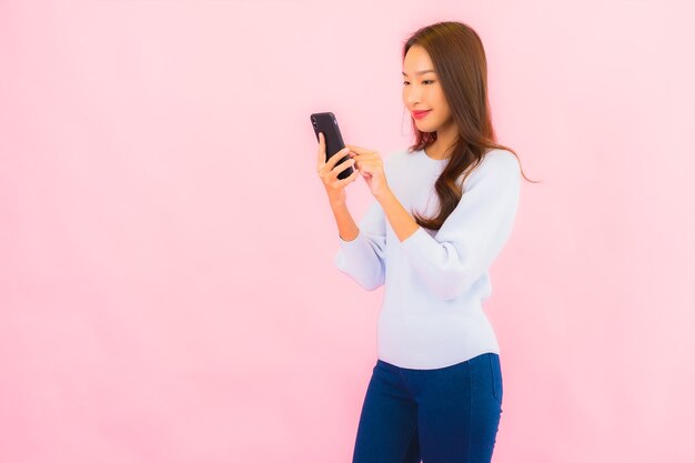 La bella giovane donna asiatica del ritratto utilizza il telefono cellulare astuto sulla parete isolata di colore rosa