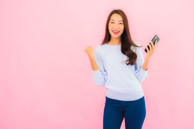 La bella giovane donna asiatica del ritratto utilizza il telefono cellulare astuto sulla parete isolata di colore rosa