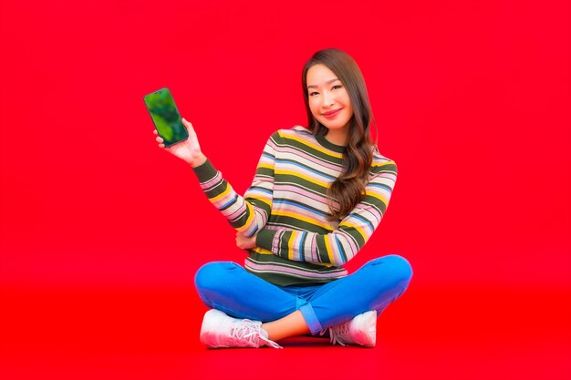 La bella giovane donna asiatica del ritratto utilizza il telefono cellulare astuto sulla parete isolata rossa