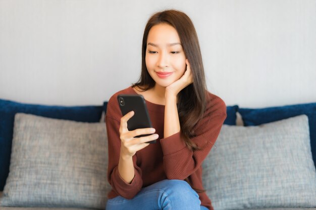 La bella giovane donna asiatica del ritratto utilizza lo smart phone sul divano nell'interno del soggiorno