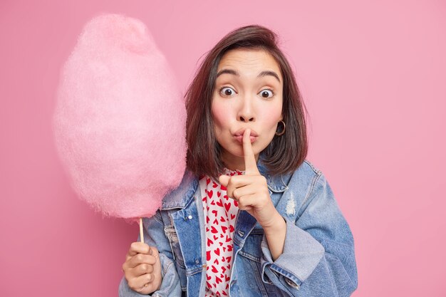 La bella giovane donna asiatica sorpresa fa il gesto del silenzio racconta che le informazioni private contengono delizioso zucchero filato vestito con pettegolezzi giacca di jeans e diffonde voci isolate sul muro rosa.