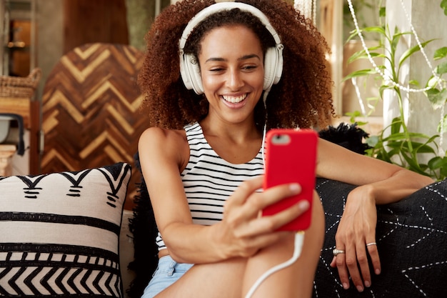 La bella giovane donna ha un'acconciatura afro, effettua una videochiamata tramite smartphone e cuffie, parla con un amico online mentre si siede al comodo divano con cuscini.