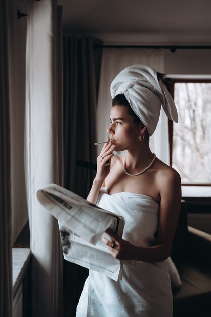 La bella giovane donna in un asciugamano fuma una sigaretta e legge il giornale