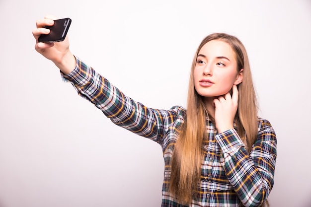 La bella giovane donna sta facendo la foto del selfie con lo smartphone isolato sulla parete bianca