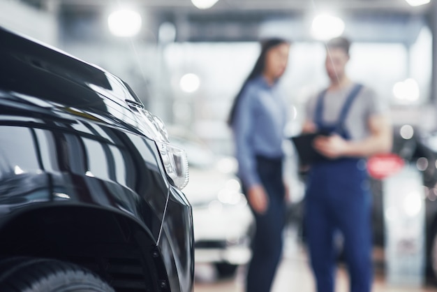 La bella giovane donna sta parlando al meccanico di automobile bello mentre ripara un'automobile in concessionaria