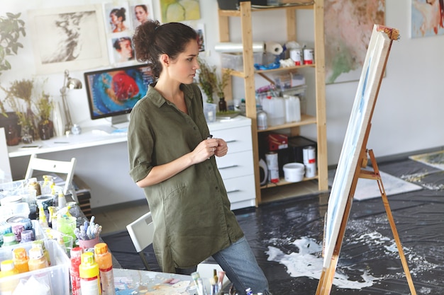 La bella giovane pittrice si è vestita casualmente in piedi di fronte al suo dipinto, studiando la sua foto con uno sguardo di valutazione, pensando a quali colori aggiungere. Concetto di arte e creatività