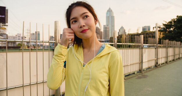 La bella giovane signora dell'atleta dell'Asia si esercita facendo uso dello smartphone per ascoltare la musica mentre corre nell'ambiente urbano. La ragazza teenager coreana che indossa gli sport copre sul ponte del passaggio pedonale nel primo mattino.