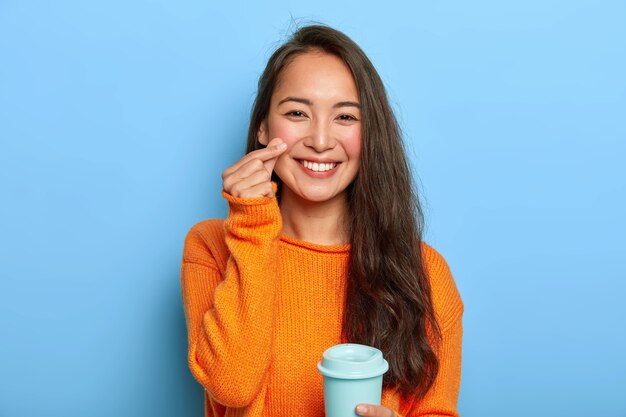 La bella mora Asain millennial donna tiene una tazza di caffè da asporto, si diverte a bere durante la pausa, indossa un caldo maglione arancione