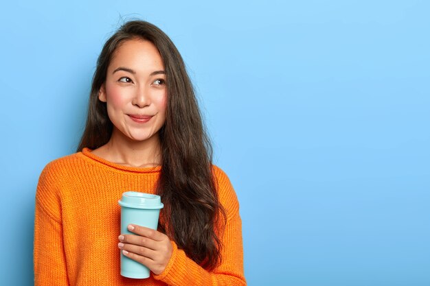 La bella mora giapponese ha i capelli lunghi, indossa un maglione caldo arancione vivido, tiene il caffè per andare