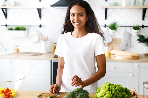 La bella mulatta sorride e tiene in mano un coltello nella moderna cucina vestita di t-shirt bianca, vicino al tavolo con verdure fresche