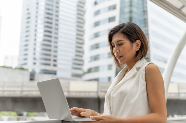 La bella ragazza asiatica che sorride in donna di affari copre facendo uso del computer portatile e dello smartphone