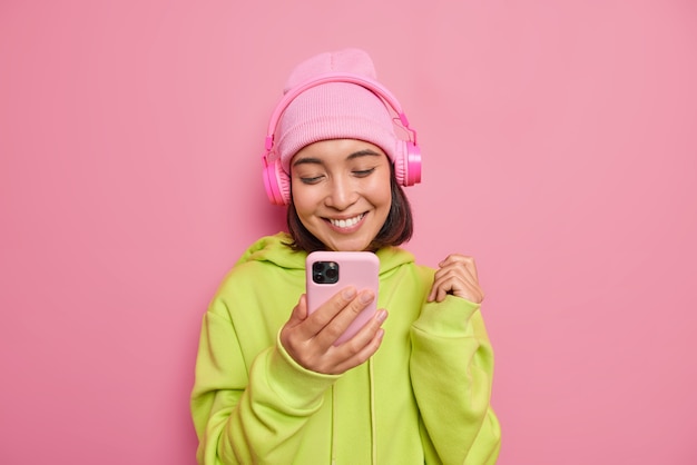 La bella ragazza asiatica felice guarda felicemente lo smartphone ascolta la musica tramite le cuffie gode della playlist preferita indossa il cappello e la felpa verde isolata sul muro rosa