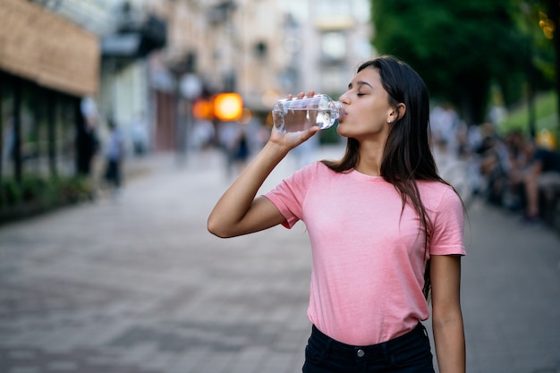 La bella ragazza beve con una bottiglia d'acqua su una strada della città