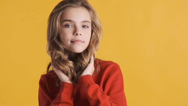 La bella ragazza bionda dell'adolescente con i capelli mossi si è vestita con un maglione rosso che posa sulla macchina fotografica isolata su fondo giallo