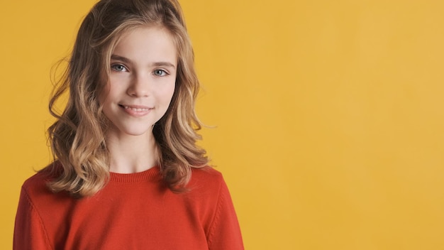 La bella ragazza bionda dell'adolescente con i capelli mossi si è vestita di maglione rosso che sorride sulla macchina fotografica sopra fondo giallo