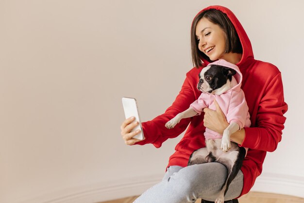 La bella ragazza caucasica prende selfie con il suo cane bulldog mentre indossa una felpa con cappuccio insieme su sfondo bianco Concetto di vera amicizia per l'animale domestico