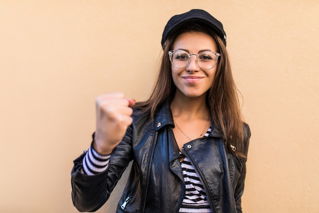 La bella ragazza di modo in giacca di pelle e cappello nero mostra il pugno forte isolato sulla parete gialla chiara