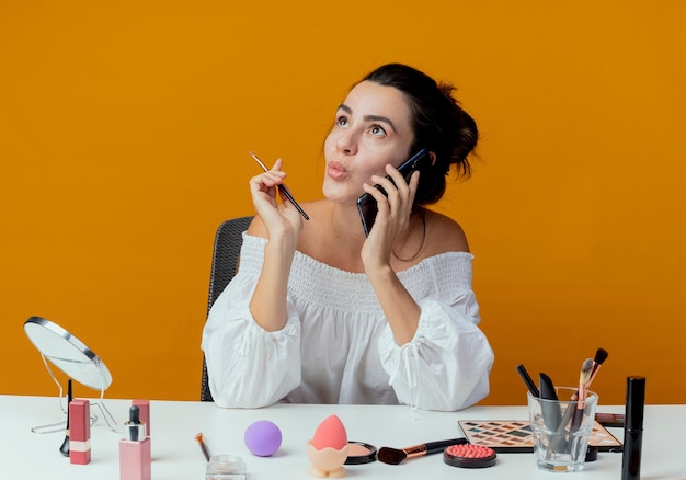 La bella ragazza emozionante si siede al tavolo con i colloqui di strumenti di trucco sul telefono che tiene la spazzola di trucco che osserva in su isolata sulla parete arancione