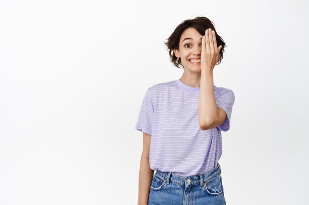 La bella ragazza felice nasconde un lato del viso, sembra eccitata, sorridente, con un occhio, in piedi in abiti casual su bianco.