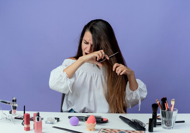 La bella ragazza infastidita si siede al tavolo con gli strumenti di trucco finge di tagliare i capelli con le forbici isolate sulla parete viola