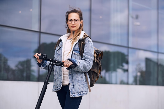 La bella ragazza pensosa con gli occhiali e con i tatuaggi sul collo e sulle mani sta guidando uno scooter elettrico.