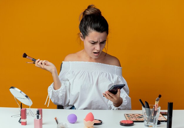 La bella ragazza scioccata si siede al tavolo con gli strumenti per il trucco tiene i pennelli per il trucco guardando il telefono isolato sulla parete arancione