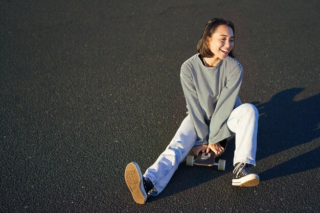 La bella ragazza teenager coreana felice si siede sul suo skateboard che gira sul longboard indossando abiti casual