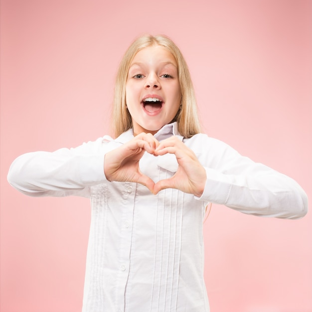 La bella ragazza teenager sorridente fa la forma di un cuore con le sue mani sui precedenti rosa. Gesto d'amore da parte di un bambino abbastanza piccolo.