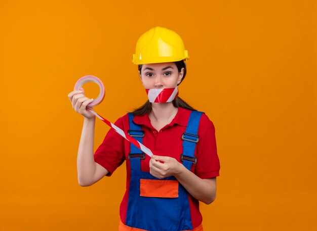 La bocca sicura della ragazza del giovane costruttore sigillata con nastro adesivo di avvertenza tiene il nastro con entrambe le mani su fondo arancio isolato