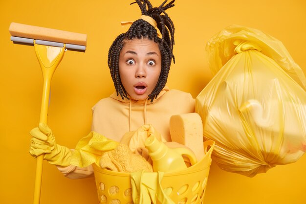 La cameriera etnica sorpresa e scioccata indossa guanti di gomma per la protezione delle mani tiene il sacchetto della spazzatura del mop