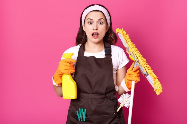 La casalinga è in preda al panico perché ha così tante cose da pulire, sta con la bocca aperta, tiene lo spray detergente e il mocio giallo in mano con guanti arancioni, ragazza scioccata sul muro rosa dello studio.