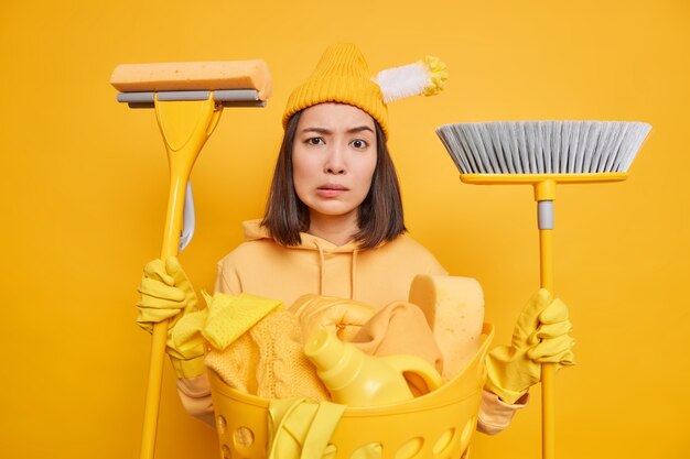 La casalinga seria perplessa non sa da cosa iniziare a pulire vestita con abiti casual usa scopa e scopa per lavare il pavimento spazza il bucato mantiene la casa pulita. Concetto di faccende domestiche