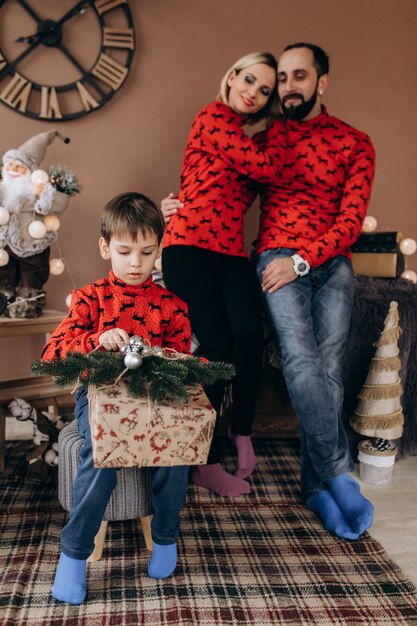 La coppia affascinante in maglioni rossi guarda il loro figlio che apre i regali prima di un albero di Natale