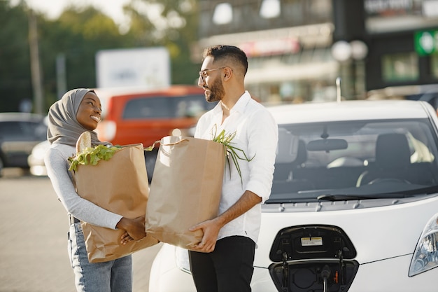 La coppia araba africana sta con generi alimentari vicino all'auto elettrica. Ricarica dell'auto elettrica alla stazione di servizio elettrica