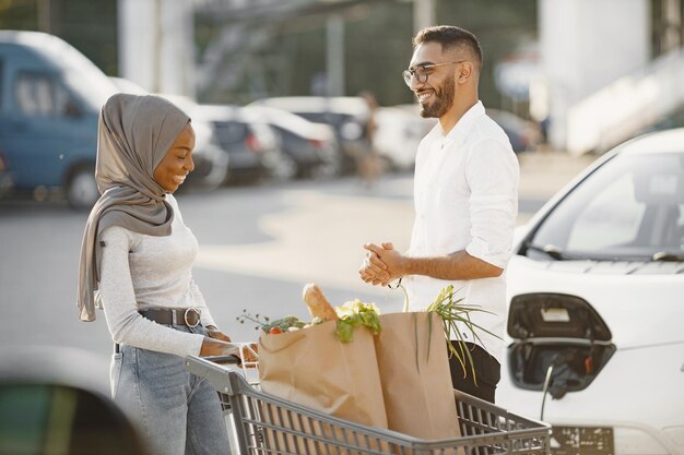 La coppia araba africana sta con generi alimentari vicino all'auto elettrica. Ricarica dell'auto elettrica alla stazione di servizio elettrica