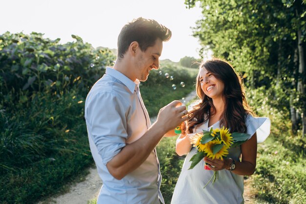 La coppia aspettante felice soffia i palloni del sapone che stanno sul campo in pieno dei girasoli gialli