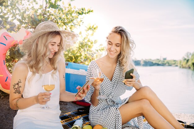 La coppia di giovani lesbiche si diverte a Riverside in una giornata di sole. Donne che trascorrono del tempo insieme sulla natura. Bere vino, fare selfie. Concetto di relazione, amore, estate, fine settimana, luna di miele.