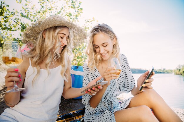 La coppia di giovani lesbiche si diverte in riva al fiume in una giornata di sole. Donne che trascorrono del tempo insieme sulla natura. Bere vino, fare selfie. Concetto di relazione, amore, estate, fine settimana, luna di miele.