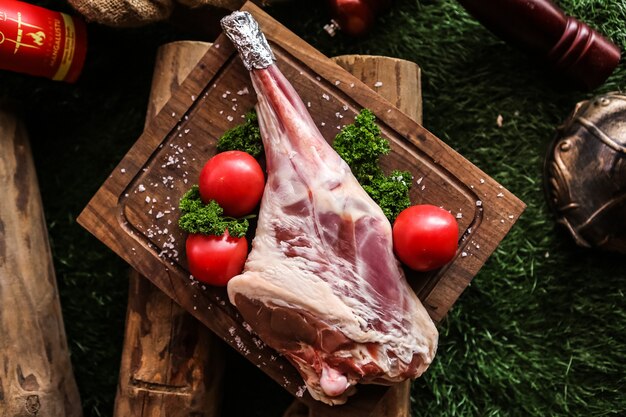 La coscia di agnello cruda sul bordo di legno ha preparato per la cottura della vista superiore di verdi di pomodoro del sale