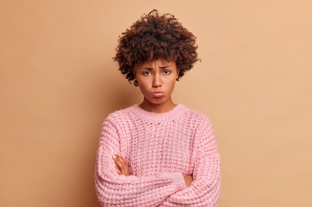 La cupa donna etnica offesa tiene le braccia conserte, porta le labbra e sembra infelice davanti, ha ferito sentimenti scopre il tradimento vestito con maglieria casual isolato su un muro marrone