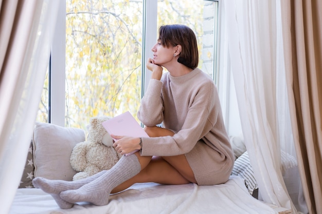 La donna a casa con il taccuino si siede sul davanzale della finestra in un maglione comodo e calzini di lana calda, fuori dalla finestra fredda fuori dalla finestra